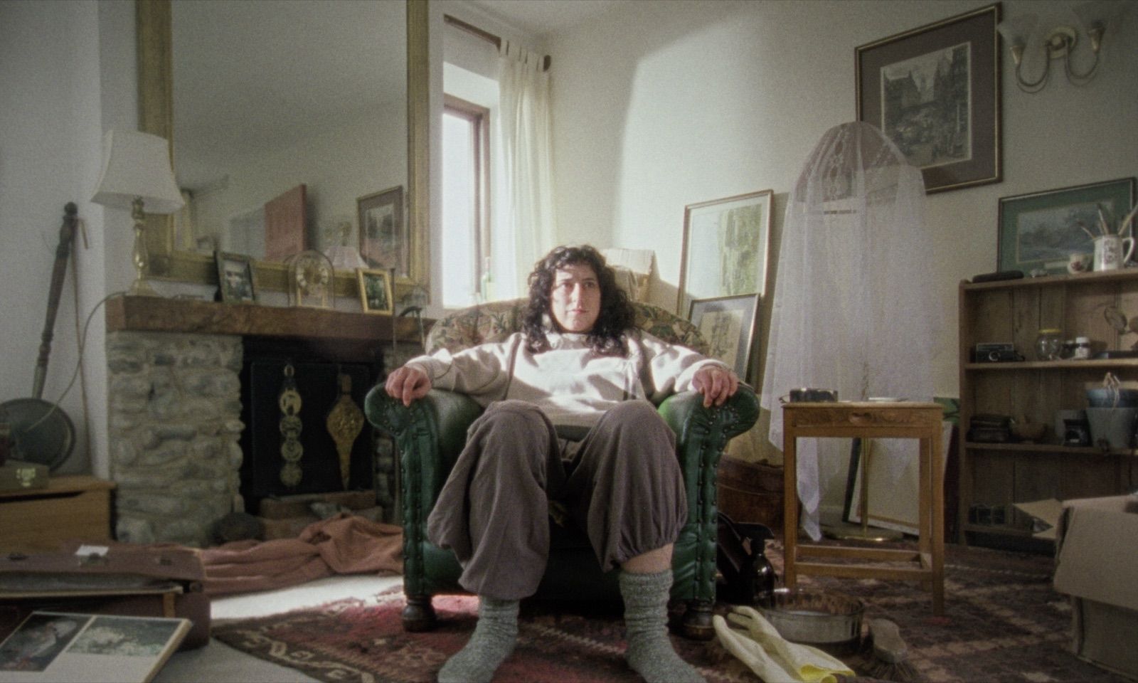 A person with dark curly hair sits squarely in a green leather armchair in a cluttered living room